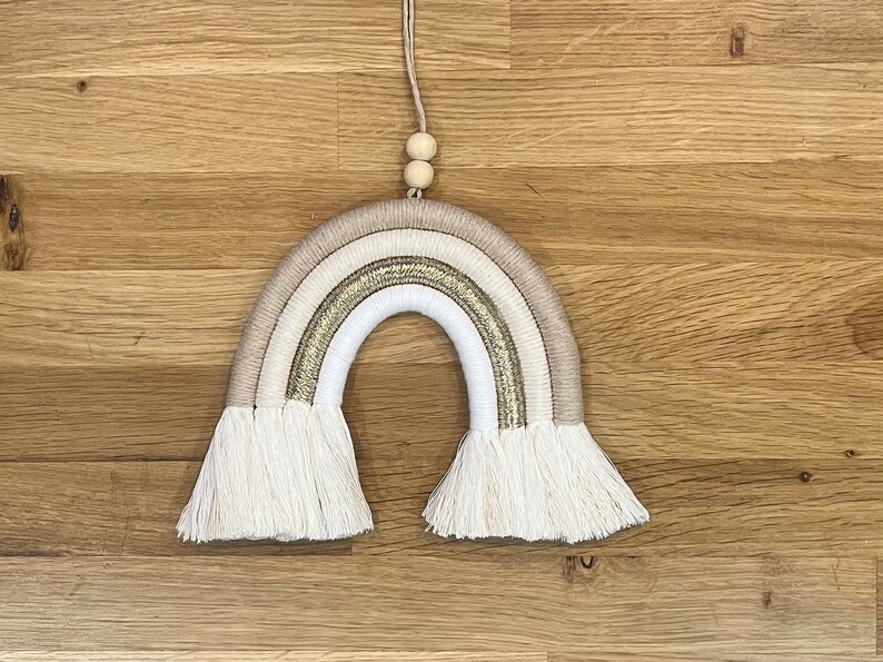 May include: A white, beige, and gold macrame rainbow wall hanging with a wooden bead hanger.