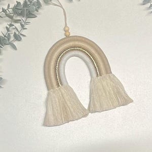 May include: A beige and white macrame rainbow wall hanging with gold thread accents and white fringe.