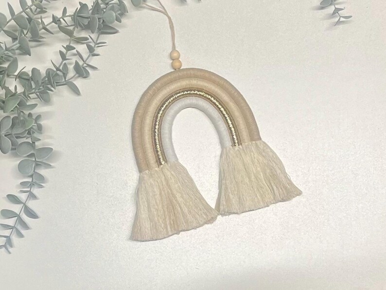 May include: A beige and white macrame rainbow wall hanging with gold thread accents and white fringe.