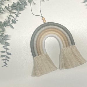 May include: A macrame rainbow wall hanging with a neutral color palette of gray, beige, and white. The rainbow has a fringe detail and is hung from a string with wooden beads.