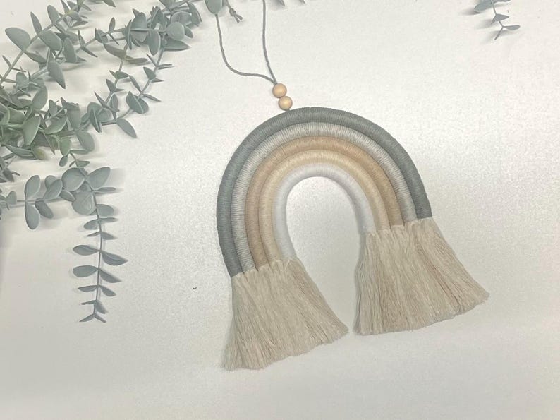 May include: A macrame rainbow wall hanging with a neutral color palette of gray, beige, and white. The rainbow has a fringe detail and is hung from a string with wooden beads.