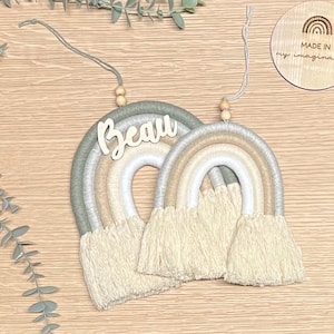 May include: Two macrame rainbows with wooden beads and a wooden name tag that says "Beau". The rainbows are in shades of green, white, and beige. The tag is attached to the larger rainbow. The smaller rainbow is hanging from the larger rainbow. The background is a light brown wooden surface.