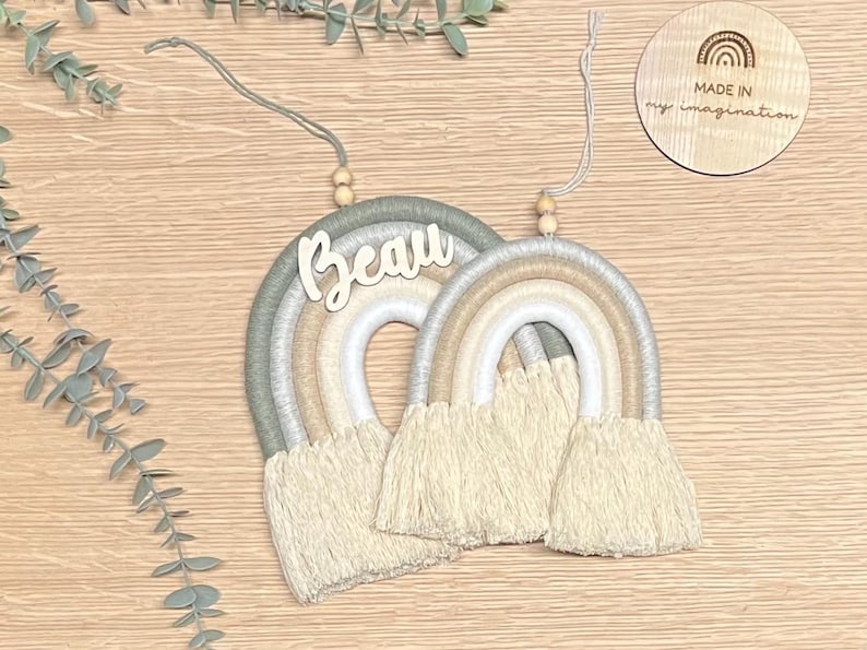 May include: Two macrame rainbows with wooden beads and a wooden name tag that says "Beau". The rainbows are in shades of green, white, and beige. The tag is attached to the larger rainbow. The smaller rainbow is hanging from the larger rainbow. The background is a light brown wooden surface.