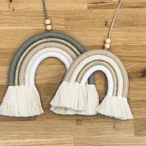 May include: Two macrame rainbows with a beige, white, and green color scheme. The rainbows are hanging from wooden beads and have white fringe.