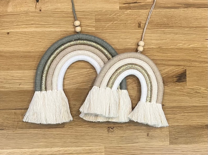 May include: Two macrame rainbows with a beige, white, and green color scheme. The rainbows are hanging from wooden beads and have white fringe.
