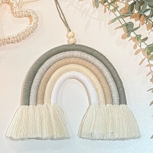 May include: A macrame rainbow wall hanging with a neutral color palette of white, beige, and green. The rainbow has a fringe at the bottom and is made with cotton cord.