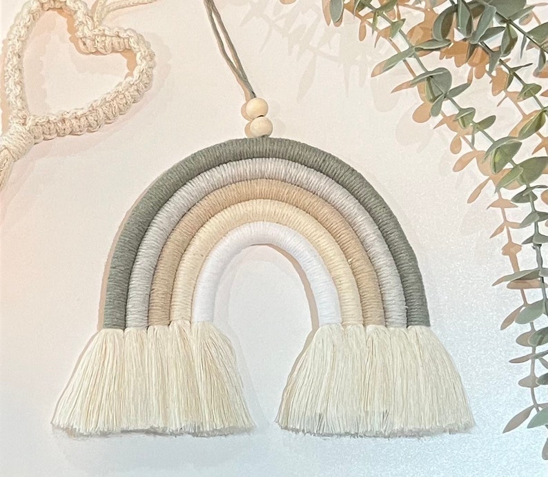 May include: A macrame rainbow wall hanging with a neutral color palette of white, beige, and green. The rainbow has a fringe at the bottom and is made with cotton cord.