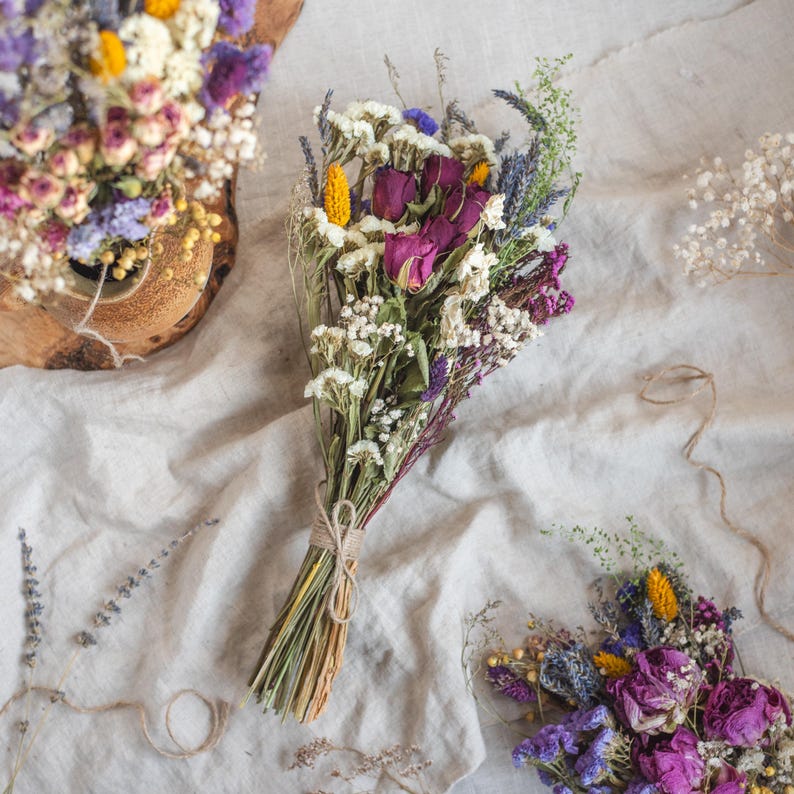 May include: A bouquet of dried flowers in shades of purple, white, and yellow. The bouquet is tied with twine and includes roses, lavender, and baby's breath.