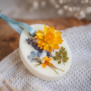 May include: A white round wax melt with a blue ribbon and dried flowers pressed into the wax. The flowers include a yellow flower, blue flowers, and green leaves.