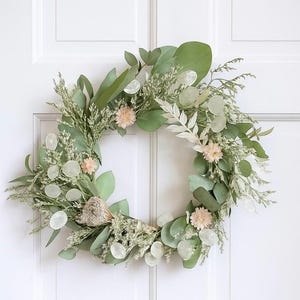 May include: A decorative wreath made from dried flowers and foliage. The wreath includes large, green eucalyptus leaves, small pink blooms, and white, coin-shaped accents. The wreath is set against a white door.
