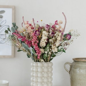 May include: A bouquet of dried flowers in shades of pink, white, and purple arranged in a white ceramic vase with a textured bubble design.