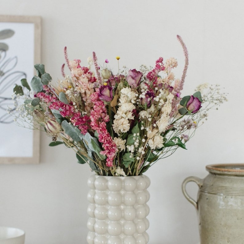 May include: A bouquet of dried flowers in shades of pink, white, and purple arranged in a white ceramic vase with a textured bubble design.