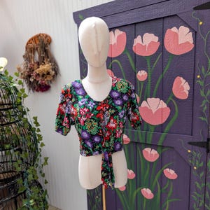 May include: A short-sleeved, tie-front top with a vibrant floral pattern in red, purple, green, and black. The top is displayed on a mannequin, showcasing its V-neck and cropped design. The background includes a painted purple door with pink flower illustrations.