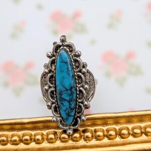 May include: A silver-toned ring featuring an oval, turquoise-colored stone with dark veining. The stone is set in an ornate, filigree-style setting. The ring is displayed on a gold-colored frame against a floral background.