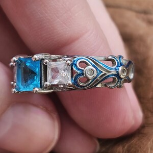 May include: A silver ring featuring three square gemstones: a vibrant blue, a clear white, and a small clear stone. The band has a blue and silver filigree design with small clear accents. The ring is a piece of jewelry.