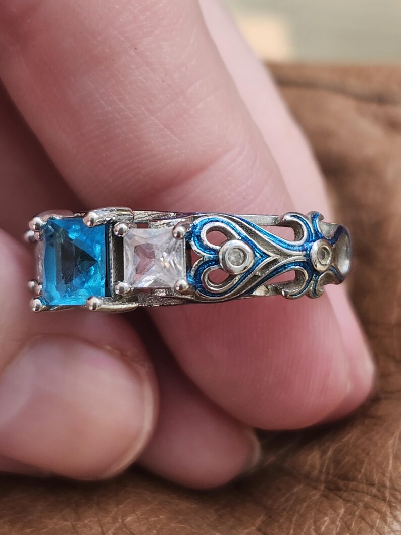 May include: A silver ring featuring three square gemstones: a vibrant blue, a clear white, and a small clear stone. The band has a blue and silver filigree design with small clear accents. The ring is a piece of jewelry.