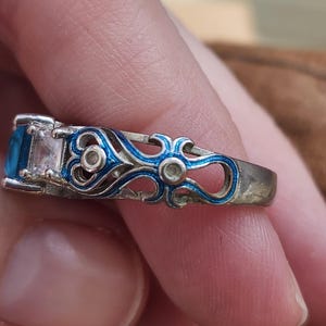May include: A silver ring with a rectangular blue gemstone and a clear rectangular gemstone. The ring features intricate blue enamel detailing along the band. The ring is held by a person.