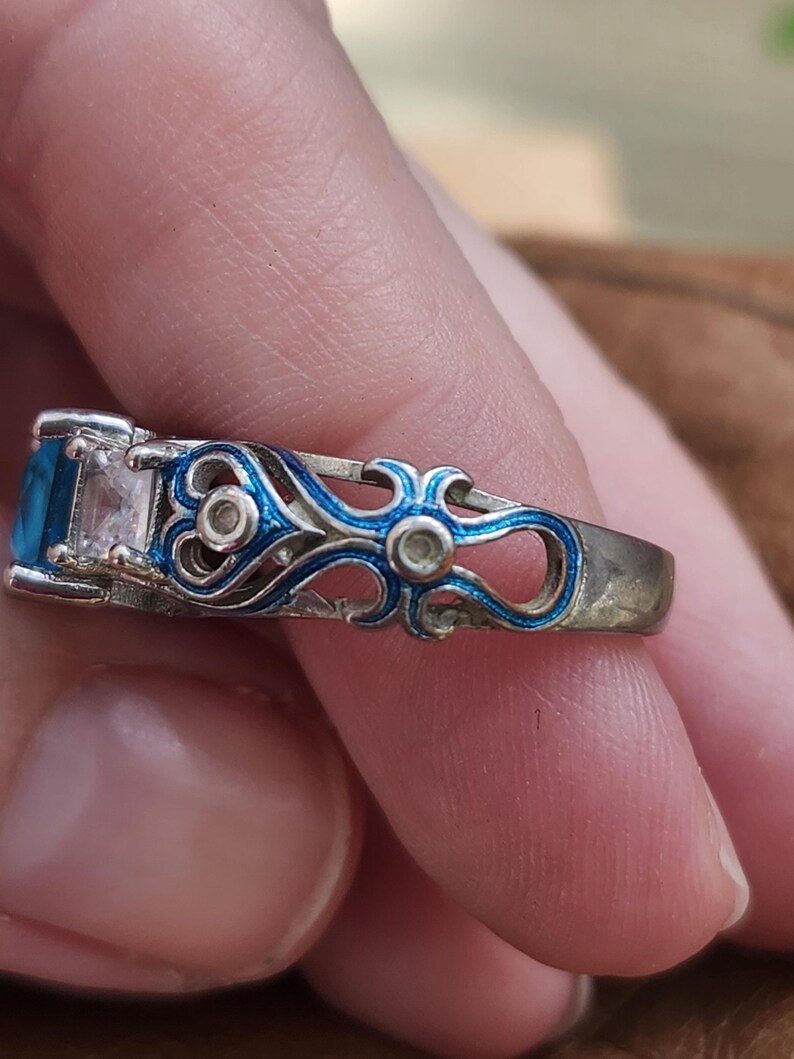 May include: A silver ring with a rectangular blue gemstone and a clear rectangular gemstone. The ring features intricate blue enamel detailing along the band. The ring is held by a person.