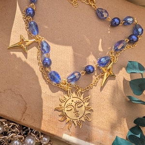 May include: A gold-toned necklace featuring a sun pendant with a serene face, accented by blue crystal beads and star charms. The necklace has a delicate chain and is displayed on a neutral surface.