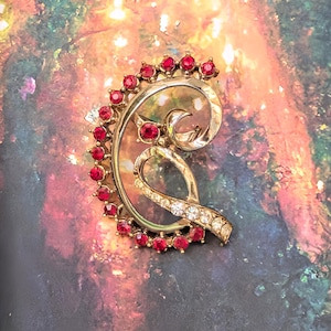 May include: A gold-tone metal brooch featuring a curved, crescent-like frame adorned with a row of small, round, vibrant red stones. The center of the brooch displays a swirling, open-work design accented with a line of clear, sparkling crystals.