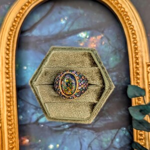 May include: A gold ring featuring a large oval-shaped dark opal center stone with a vibrant play of color. The band is encrusted with numerous small, multi-colored gemstones in shades of pink, blue, green, and yellow, set in a gold metal frame.