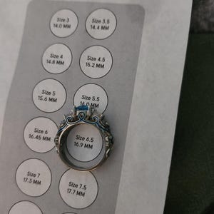 May include: A silver ring with a blue gemstone and intricate detailing. The ring is placed on a ring size chart, with a size 6.5 (16.9 mm) visible. The chart displays various ring sizes and their corresponding millimeter measurements.
