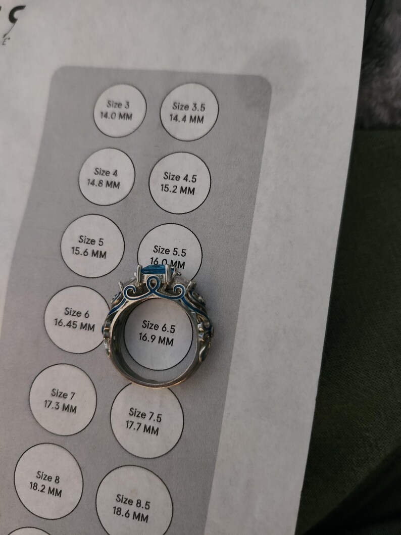 May include: A silver ring with a blue gemstone and intricate detailing. The ring is placed on a ring size chart, with a size 6.5 (16.9 mm) visible. The chart displays various ring sizes and their corresponding millimeter measurements.