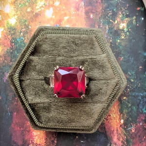 May include: A ring featuring a large, square-cut, vibrant red gemstone set in a metallic band. The ring is displayed in a hexagonal, olive green velvet box. The stone has a faceted surface that reflects light, and the setting includes small clear side stones.