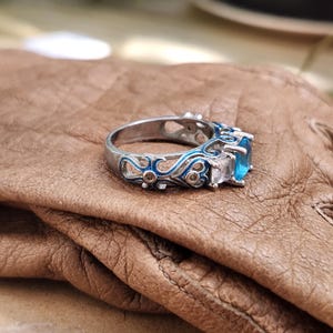 May include: A silver ring with a blue gemstone and white accent stones. The ring features intricate blue enamel detailing along the band. The ring is set on a brown leather surface.