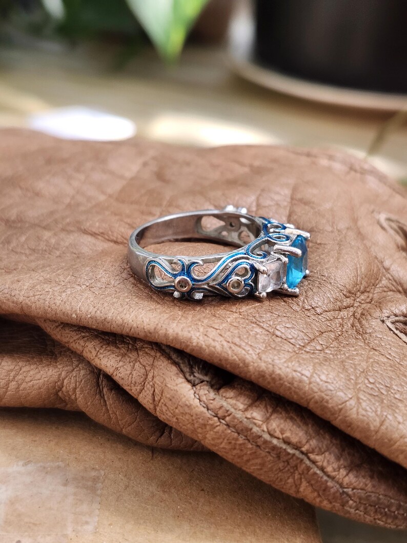 May include: A silver ring with a blue gemstone and white accent stones. The ring features intricate blue enamel detailing along the band. The ring is set on a brown leather surface.