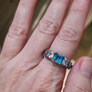 May include: Silver ring featuring a square, light blue gemstone flanked by two clear, square-cut stones. The band has an ornate, openwork design. The ring is worn on a finger, showcasing the jewelry's details and color.
