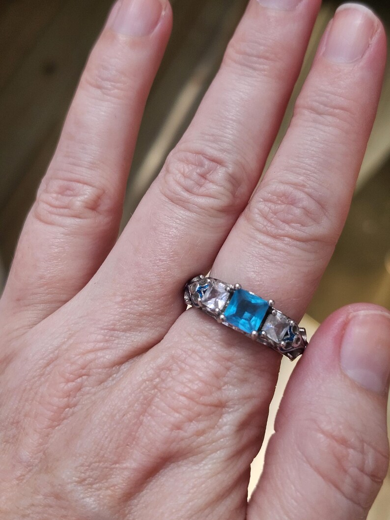 May include: Silver ring featuring a square, light blue gemstone flanked by two clear, square-cut stones. The band has an ornate, openwork design. The ring is worn on a finger, showcasing the jewelry's details and color.