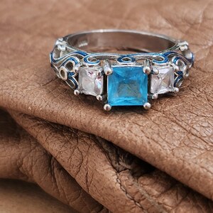 May include: A silver ring with a square, light blue gemstone in the center, flanked by two smaller, square, clear gemstones. The ring has intricate blue enamel detailing on the band, creating a decorative and elegant design.