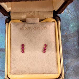 May include: A pair of gold earrings with three red gemstones in each, displayed in a vintage brown velvet jewelry box. The box is open, revealing the earrings against a beige background. The box is labeled "14 KT. GOLD".