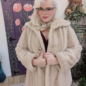 Puede incluir: Un abrigo de piel sintética beige con capucha y botones grandes. El abrigo se lleva sobre un vestido burdeos oscuro. El fondo incluye una puerta morada con detalles florales y una jaula decorativa.