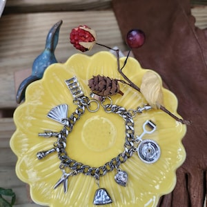 May include: A silver charm bracelet with various charms, including a fan, scissors, and a bell, rests on a yellow, flower-shaped dish. The bracelet is displayed with decorative elements like berries and a brown leather glove.