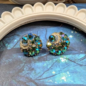 May include: A pair of round clip-on earrings featuring a cluster of iridescent blue and green faceted rhinestones set in a gold-tone metal frame. Each earring includes a small, textured gold-tone leaf or feather accent integrated into the circular design.