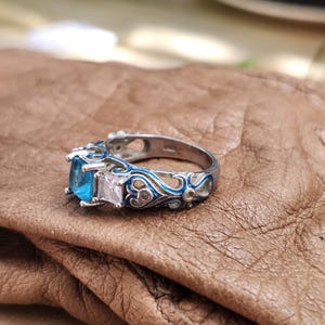 May include: A silver ring with a square, light blue gemstone and a smaller, square, clear gemstone. The ring has intricate blue enamel detailing and is set on a brown surface. The ring is a piece of jewelry.
