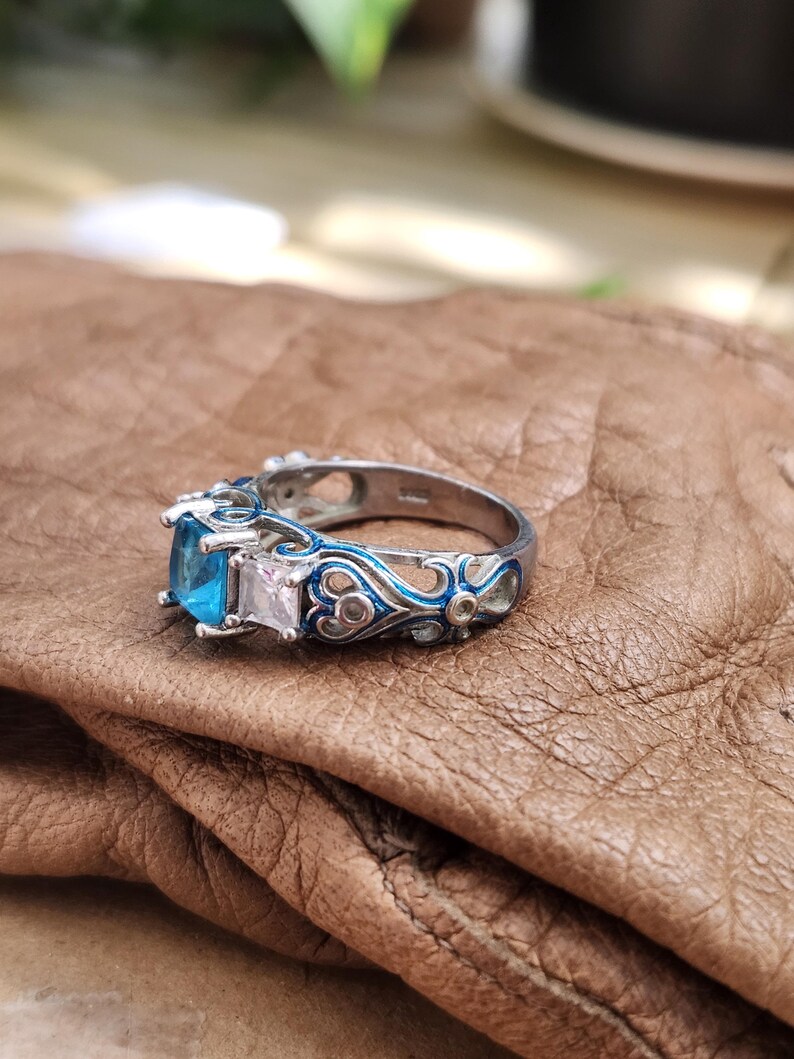 May include: A silver ring with a square, light blue gemstone and a smaller, square, clear gemstone. The ring has intricate blue enamel detailing and is set on a brown surface. The ring is a piece of jewelry.