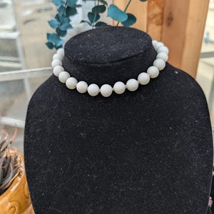 May include: A white beaded necklace displayed on a black velvet bust. The necklace features large, round beads strung together. The background includes a window and decorative greenery, suggesting a jewelry or accessory theme.