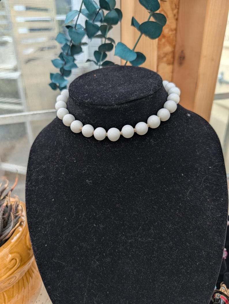 May include: A white beaded necklace displayed on a black velvet bust. The necklace features large, round beads strung together. The background includes a window and decorative greenery, suggesting a jewelry or accessory theme.