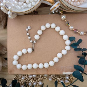 May include: A collection of vintage jewelry, including a white beaded necklace with a silver clasp, pearl necklaces, and gold chains. The necklaces are arranged with teacups and decorative greenery.