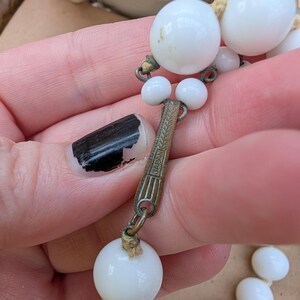 May include: Close-up of a vintage white beaded necklace. The necklace features large, round beads and smaller accent beads. The clasp is metal with the word "JAPAN" engraved on it. The beads are strung on a beige cord.