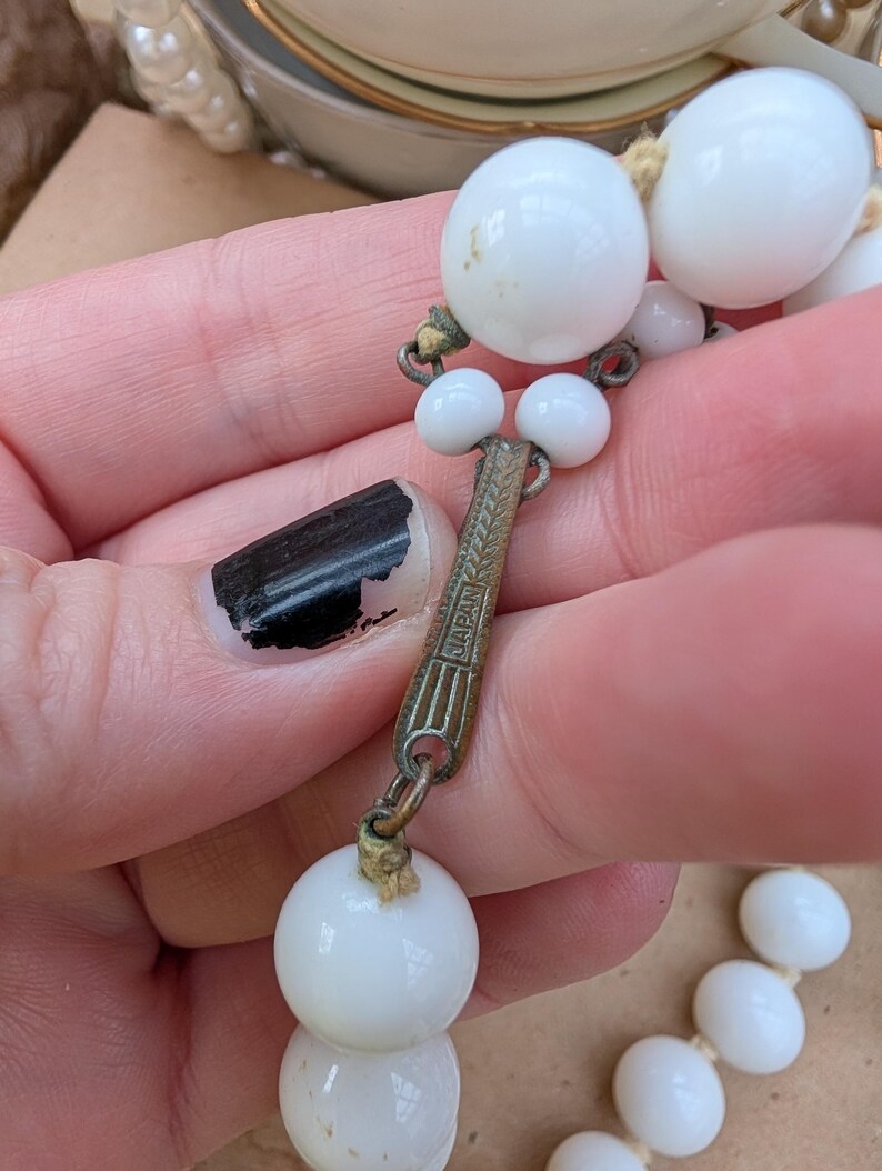 May include: Close-up of a vintage white beaded necklace. The necklace features large, round beads and smaller accent beads. The clasp is metal with the word "JAPAN" engraved on it. The beads are strung on a beige cord.
