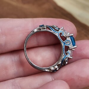 May include: A silver ring with a large, square, blue gemstone. The ring features intricate blue enamel detailing around the gemstone and along the band, creating a decorative and ornate design. The ring is held in a hand.