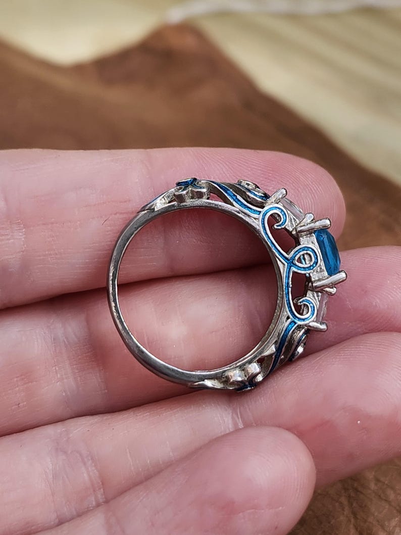 May include: A silver ring with a large, square, blue gemstone. The ring features intricate blue enamel detailing around the gemstone and along the band, creating a decorative and ornate design. The ring is held in a hand.