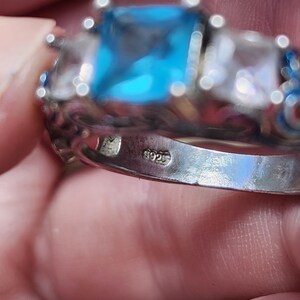 May include: A silver ring with a large, square, light blue gemstone as the centerpiece, flanked by two smaller, clear gemstones. The ring has intricate detailing and a 925 stamp.