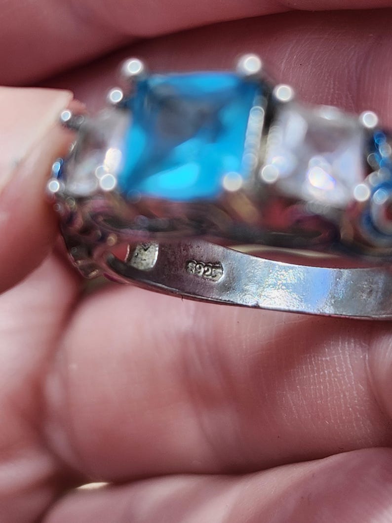 May include: A silver ring with a large, square, light blue gemstone as the centerpiece, flanked by two smaller, clear gemstones. The ring has intricate detailing and a 925 stamp.