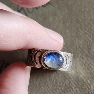 May include: A silver ring with a moonstone cabochon and three crescent moon engravings. The ring is worn on a finger.