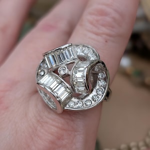 May include: A silver-toned ring with a cluster design. The ring features multiple rectangular and round-cut clear gemstones. The ring is set on a finger, with the background out of focus.
